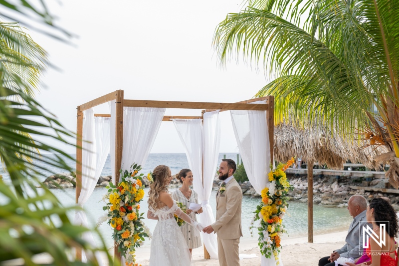 A Beach Wedding Ceremony Takes Place Under a Floral Arch Adorned With Yellow and Orange Blooms, Surrounded by Palm Trees and Tranquil Waters at a Tropical Location