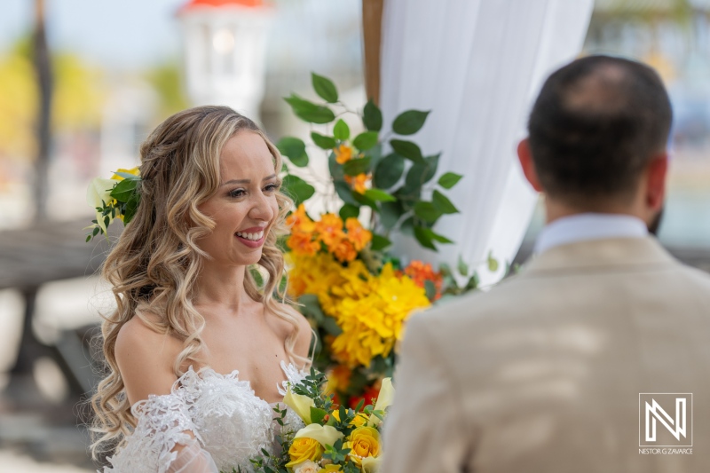 A Bride Smiles Joyfully While Exchanging Vows With Her Partner During an Outdoor Wedding Ceremony Surrounded by Vibrant Flowers on a Sunny Day