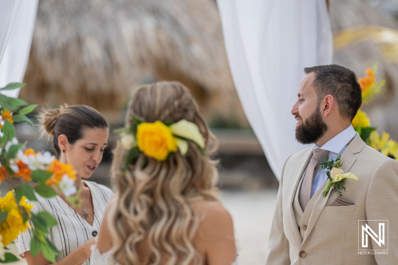 A Beach Wedding Ceremony Takes Place at Sunset With Guests Gathered and Floral Arrangements Surrounding the Couple, While an Officiant Reads Vows in a Tropical Setting