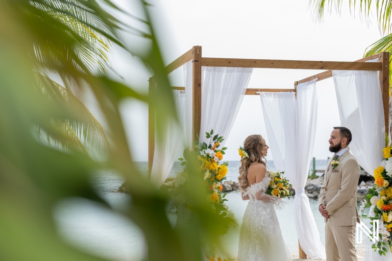 A Couple Exchanges Vows Under a Floral Arch on a Beach During a Romantic Outdoor Wedding Ceremony Surrounded by Tropical Scenery