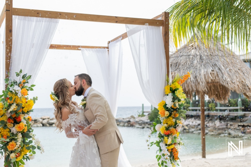 Romantic Beach Wedding Ceremony With a Couple Kissing Under a Decorated Arch Adorned With Vibrant Flowers at a Scenic Coastal Location During Daylight