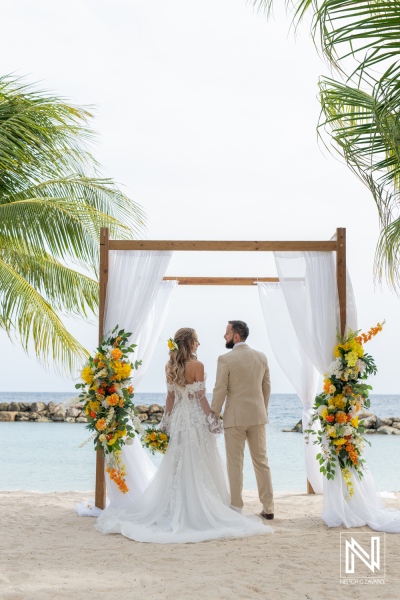 A Couple Exchanges Vows Under a Floral Arch on a Sunny Beach, Surrounded by Palm Trees and the Ocean, Celebrating Their Wedding in a Picturesque Location