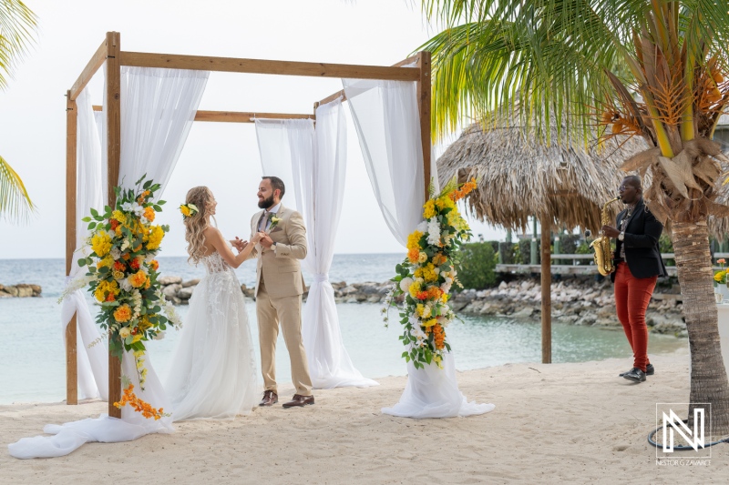 A Romantic Beach Wedding Ceremony Takes Place at a Tropical Location, Featuring a Couple Exchanging Vows Under a Floral Arch With Palm Trees and Ocean Waves in the Background