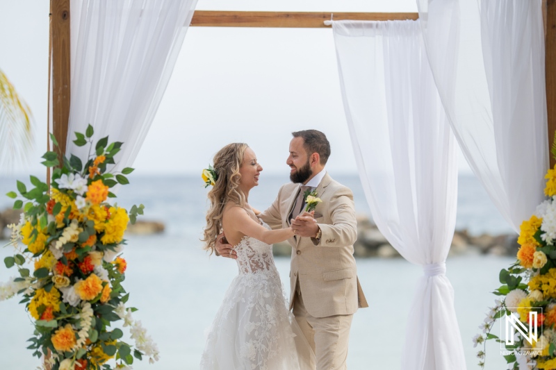 A Romantic Beach Wedding Ceremony Between a Couple Under a Floral Arch, With Vivid Flowers Framing the Serene Ocean View in the Background During a Sunny Day