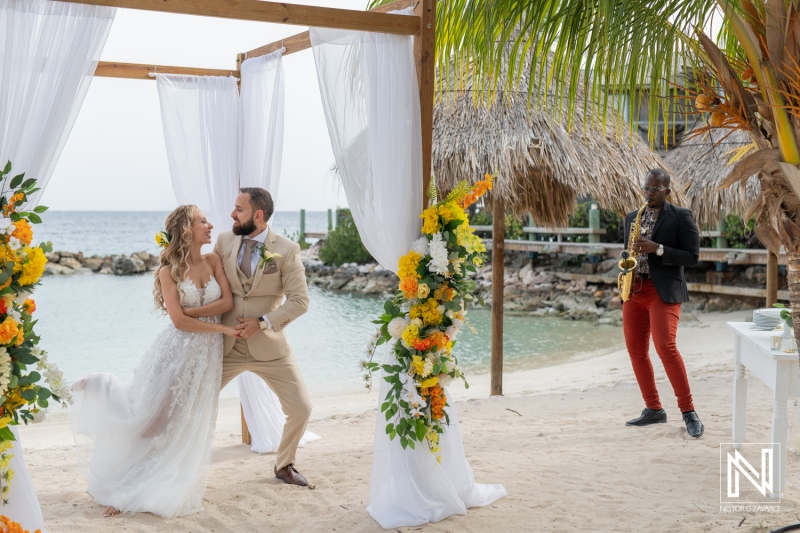 A Beautiful Beach Wedding Ceremony Unfolds With a Couple Exchanging Vows, Adorned With Tropical Flowers While a Saxophonist Plays Nearby During Sunset