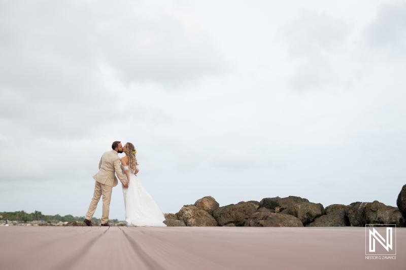 A Couple Shares a Romantic Kiss on a Coastal Walkway, Dressed in Wedding Attire, Surrounded by Rocks and a Cloudy Sky in a Serene Outdoor Setting During Their Wedding Celebration