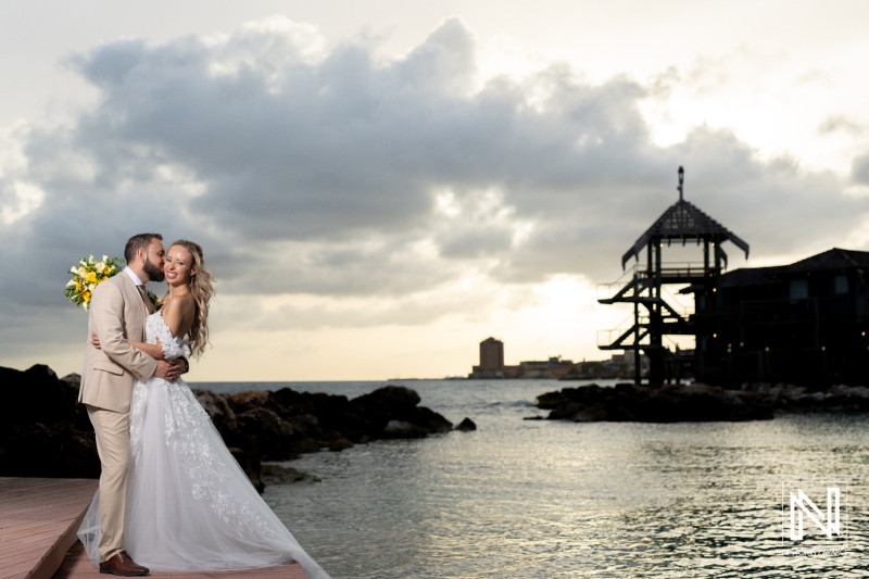 Couple Shares a Romantic Moment by the Seaside at Sunset, Celebrating Their Wedding Amidst Tranquil Waters and a Beautiful Sky