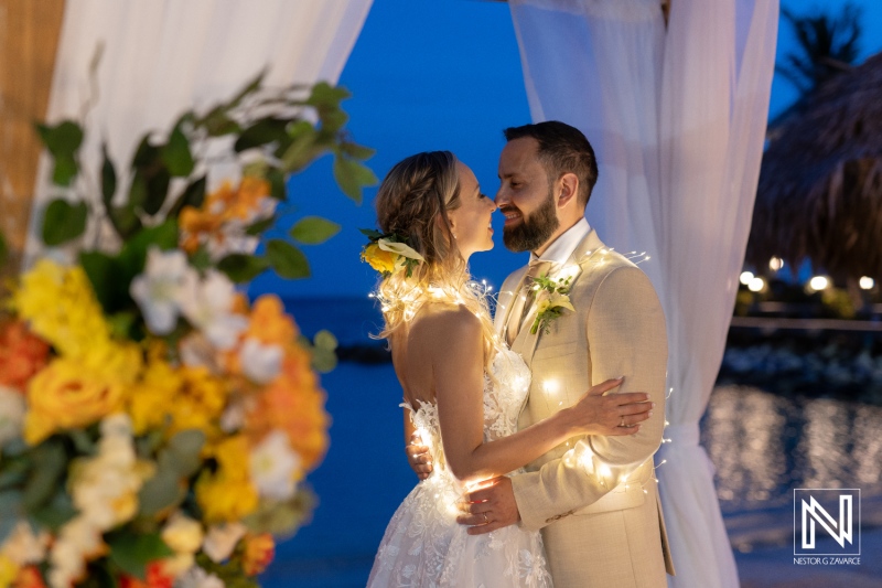 Couple Celebrating Their Wedding in a Tropical Setting at Sunset, Wrapped in String Lights With Floral Decorations Surrounding Them