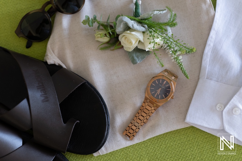 Casual summer outfit laid out on a green surface featuring stylish sandals, a wooden watch, a white shirt, and a small floral arrangement, perfect for a relaxed day out