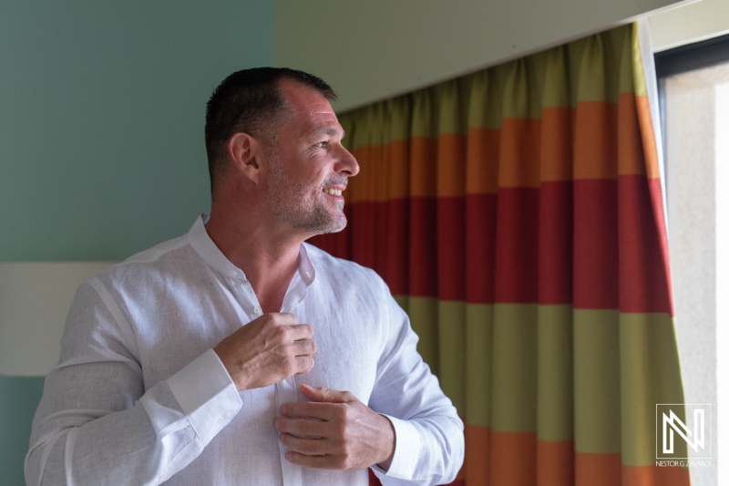 A man adjusts his shirt while standing near a window in a brightly colored room, capturing a moment of preparation in the morning light