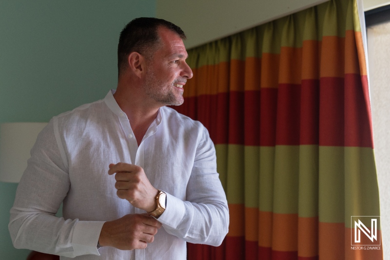 A man adjusts his shirt while looking out the window in a brightly lit room with colorful curtains during the afternoon