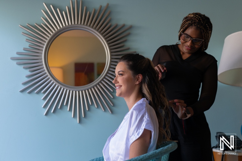 A woman prepares for a special event while getting her hair styled in a bright, modern room with a sunburst mirror and a stylish lamp in the background