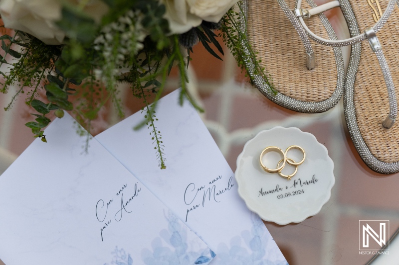 Elegant wedding details featuring personalized invitations, rings, and sandals at an outdoor venue in the afternoon sun