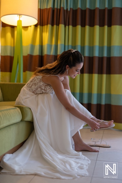 A woman in a wedding dress lounges on a green sofa while putting on her shoes in a brightly decorated room before the ceremony begins in the afternoon light