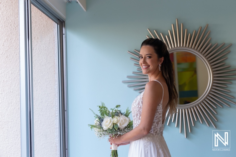 A bride stands near a sunburst mirror holding a bouquet of white roses, smiling brightly as she prepares for her wedding in a stylish indoor setting