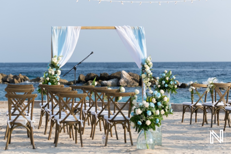 Beach wedding ceremony setup with floral decor and chairs arranged near the ocean under a clear sky in a tropical location