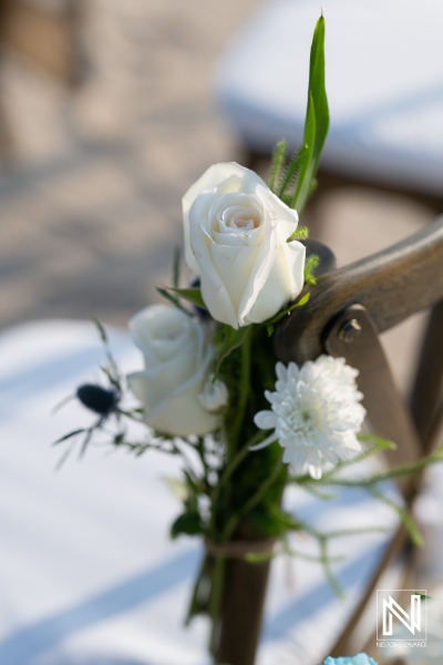 Elegant white flowers adorn wooden chairs at an outdoor ceremony in a serene setting during a sunny day