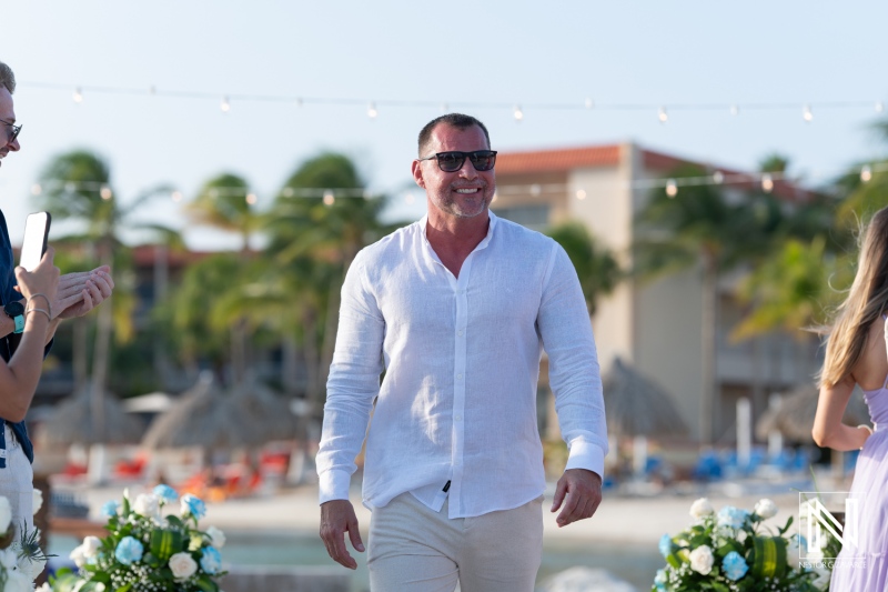 A joyful celebration unfolds at a beachfront wedding as the groom arrives in a white shirt and sunglasses during a sunny afternoon