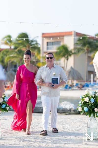 A couple walks together along the beach during a wedding ceremony at a tropical resort in the afternoon sunlight, surrounded by flowers and palm trees