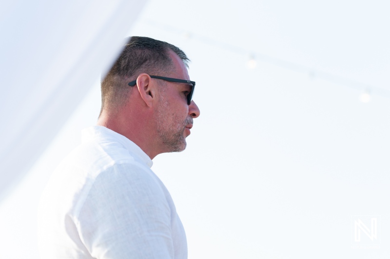 A man in sunglasses stands against a bright background during a daytime event, showcasing a stylish and confident look while enjoying the sunny atmosphere