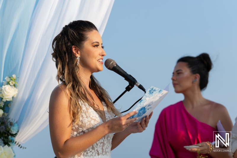 A woman delivers heartfelt vows at a beach wedding ceremony under a blue sky, while a guest stands attentively near her, capturing a moment of love and celebration