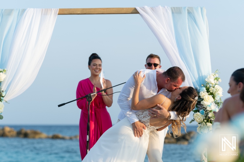 A couple celebrates their wedding with a romantic kiss at a beachfront ceremony during sunset, attended by friends and family in a tropical paradise