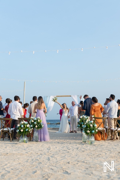 A joyful beach wedding ceremony at sunset with guests celebrating the couple and a beautiful floral arch in the background