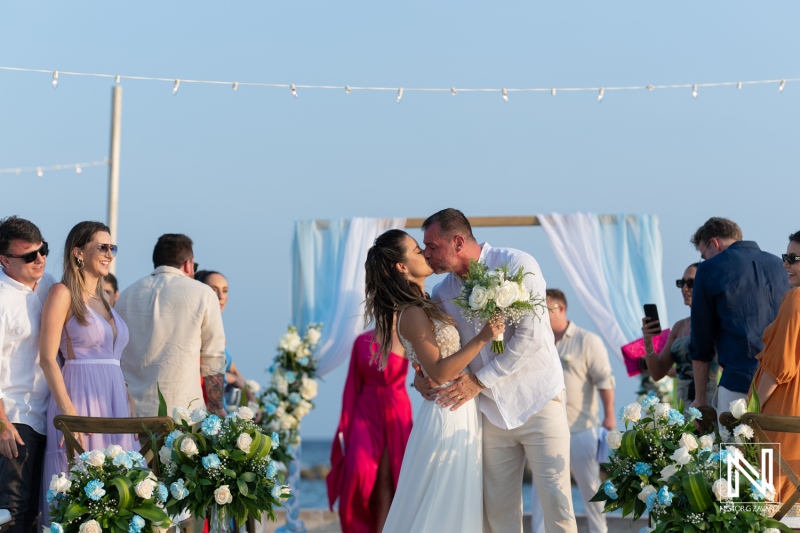 Couple shares a romantic kiss under a beautiful sky during their beach wedding ceremony surrounded by friends and family in a tropical location