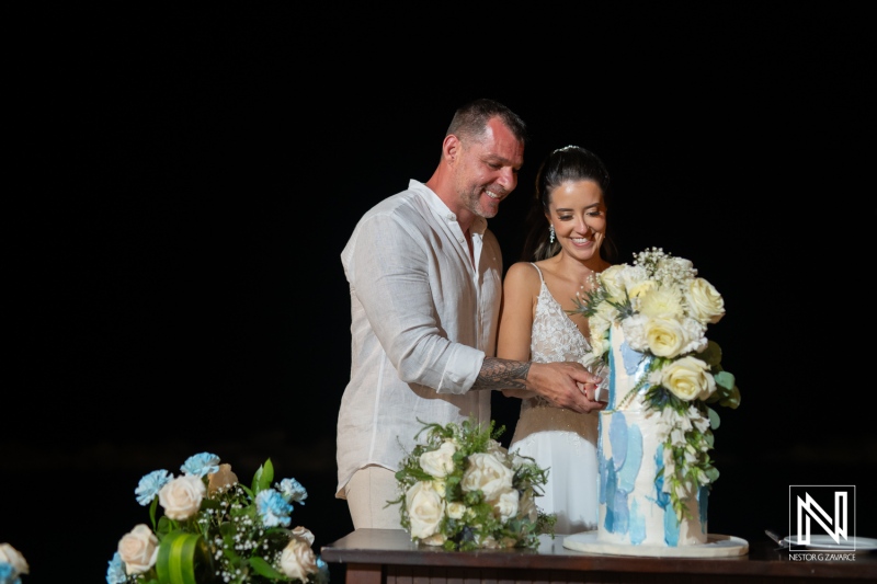 Couple cutting a beautiful wedding cake adorned with white and blue flowers on their special day at a beachside venue during the evening