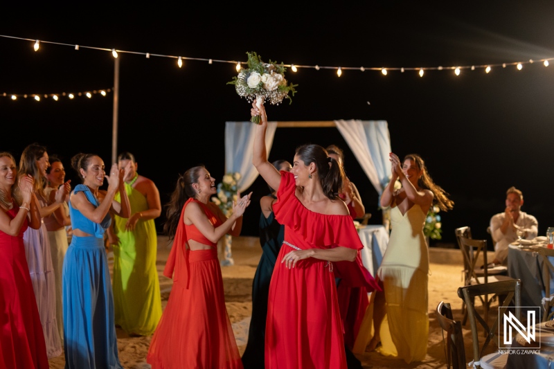 A joyful bridal party celebrates under twinkling lights, showcasing colorful dresses while a bride-to-be holds her bouquet high during nighttime festivities by the beach