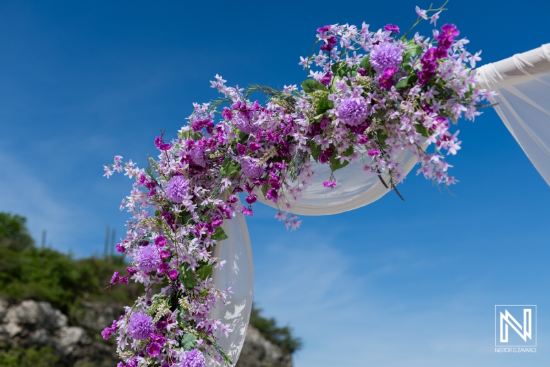 Beautiful Floral Arch Adorned With Purple Blooms and Delicate White Fabric at a Sunny Outdoor Wedding Venue