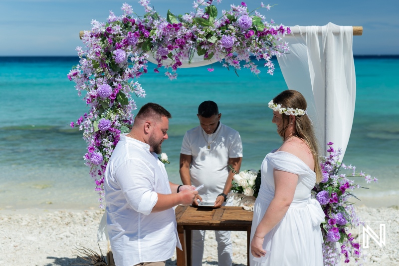 A Couple Exchanges Vows During a Beachfront Wedding Ceremony Decorated With Purple Flowers, Showcasing a Beautiful Ocean Backdrop Under Clear Skies