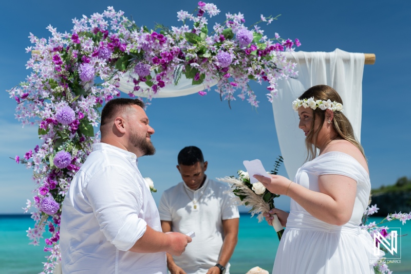 A Serene Beach Wedding Ceremony With a Floral Arch, Featuring a Bride and Groom Exchanging Vows Under a Bright Blue Sky in a Tropical Location