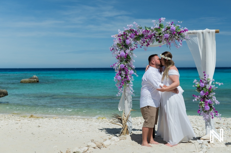 Couple Sharing a Romantic Kiss Under a Floral Arch on a Serene Beach During a Sunny Day, With Turquoise Waters and a Clear Blue Sky in the Background