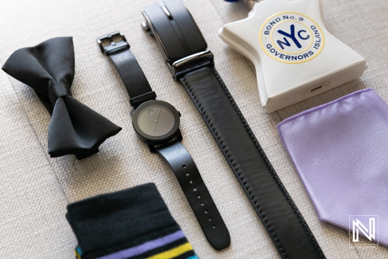 A Stylish Assortment of Accessories Laid out on a Textured Surface Featuring a Bow Tie, Watch, Leather Strap, and a Fragrance Bottle From Governors Island, Showcasing Elegance and Attention to Detail