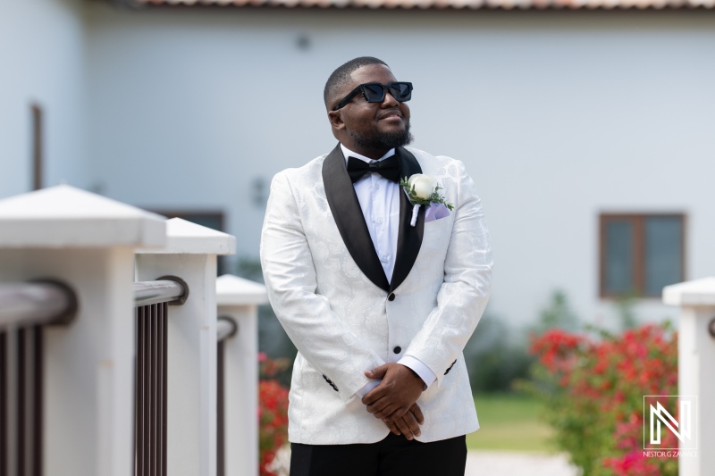 A Groom in a Stylish White Tuxedo and Sunglasses Poses Confidently in a Garden Setting Prior to His Wedding Ceremony at a Beautiful Venue