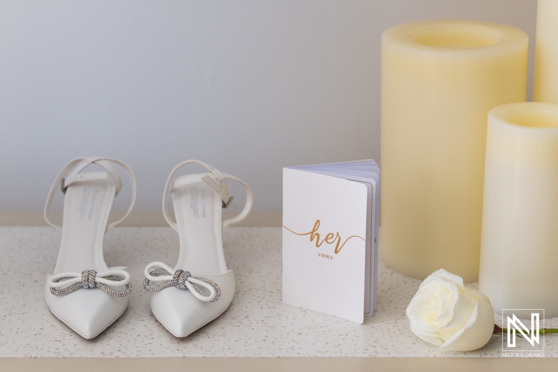 Elegant White Bridal Shoes With Decorative Bows Positioned Beside a Charming Wedding Vow Book and Soft Candles, Creating a Romantic Atmosphere for a Wedding Day