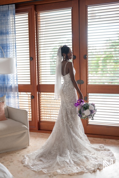 A Bride in a Beautiful Lace Gown Holds a Bouquet While Standing by Large Windows Showcasing Natural Light in an Elegant Interior Setting