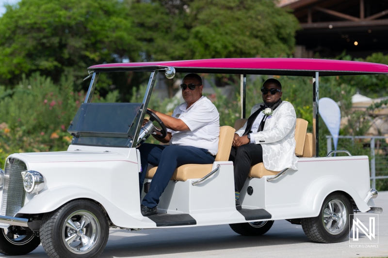 A Couple Enjoys a Sunny Day Riding in a Vintage-Style White Golf Cart With a Pink Canopy at an Outdoor Event Location Surrounded by Lush Greenery