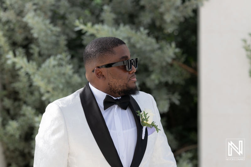 A Well-Dressed Man in a White Suit With a Bow Tie and Sunglasses Stands Confidently Outdoors Surrounded by Greenery, Enjoying a Vibrant Afternoon