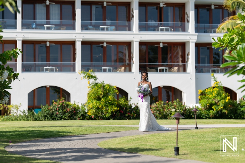 A Bride in a Stunning White Gown Holds a Bouquet While Walking Through a Beautiful Garden Outside a Resort During a Sunny Day