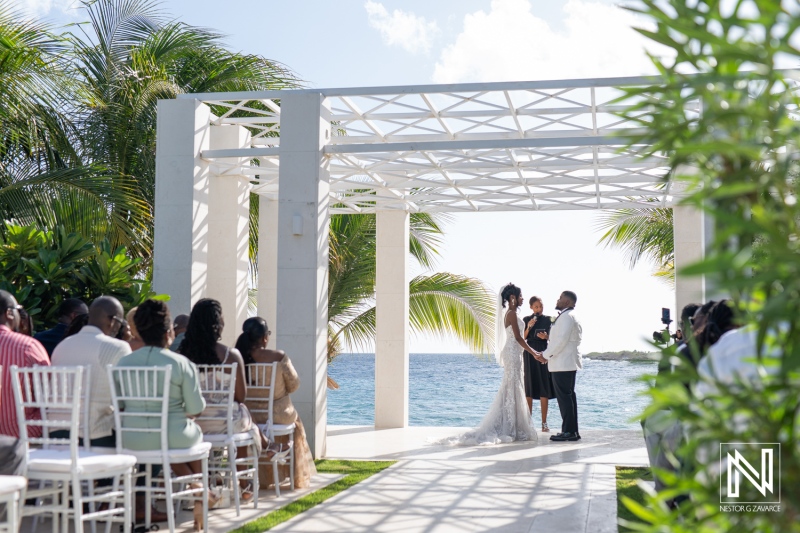 A Wedding Ceremony by the Ocean Featuring a Bride, Groom, and Guests Gathered Under a Trellis in a Tropical Setting, Surrounded by Palm Trees on a Sunny Day