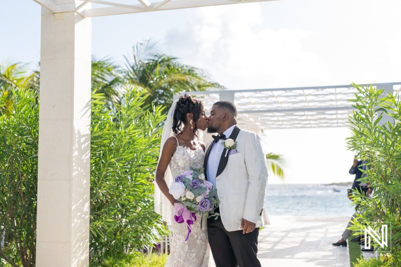 A Joyful Couple Shares a Kiss During Their Beachside Wedding Ceremony Under the Clear Sky and Palm Trees, Celebrating Their Love With Family and Friends