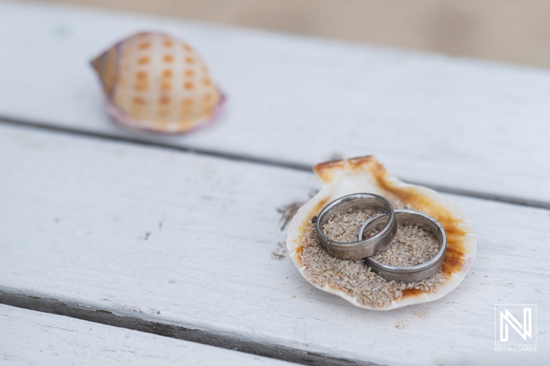 Two wedding rings resting on a shell filled with sand, surrounded by a calm beach setting during a sunny day