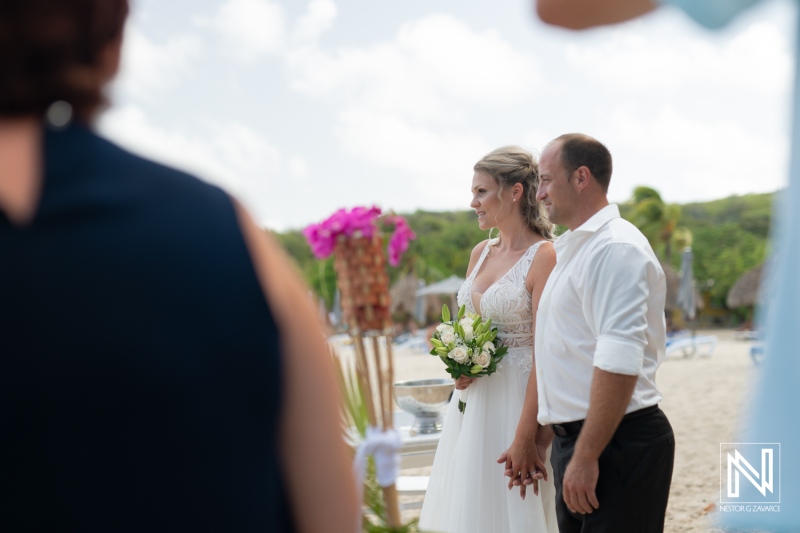 Couple exchanging vows during a beach wedding ceremony at sunset with floral arrangements and guests in attendance