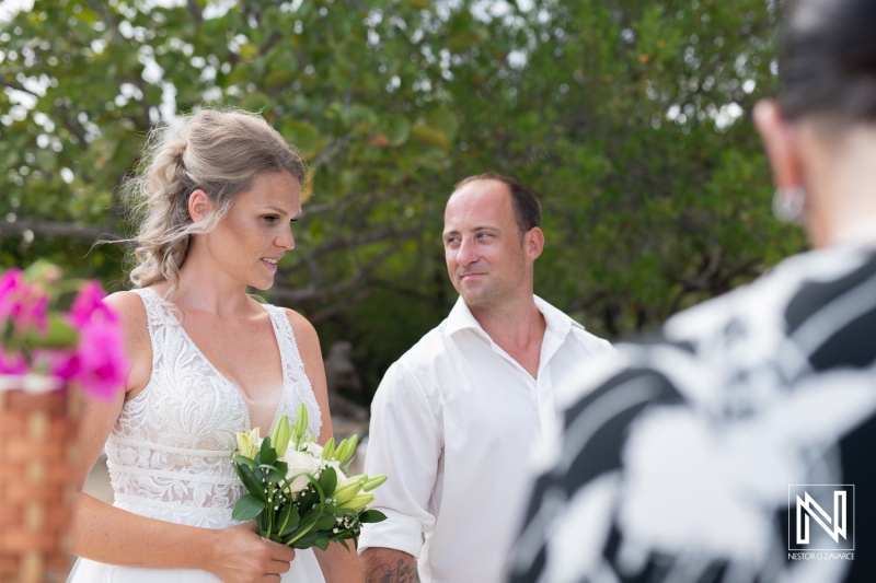 A couple stands together at a wedding ceremony outdoors, surrounded by greenery, as the bride smiles and holds a bouquet of flowers