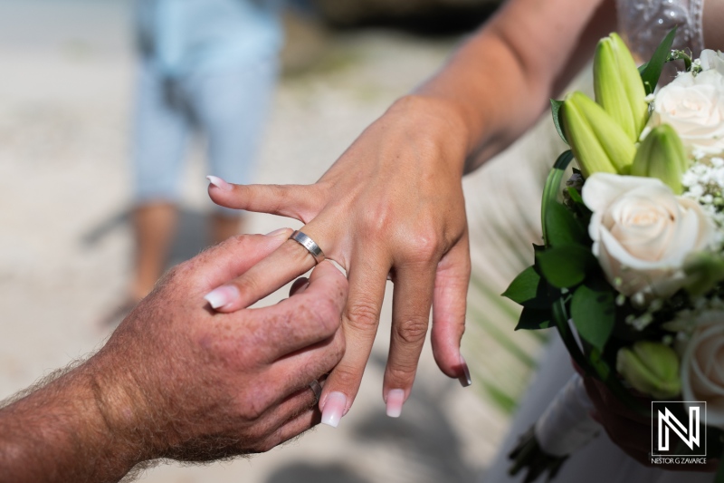 A wedding ceremony on a beach, where a groom places a wedding ring on the bride\'s finger while she holds a bouquet of flowers under the warm sunlight