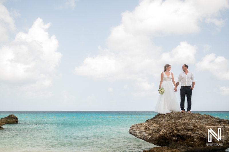 A couple in wedding attire holds hands while standing on a rocky outcrop overlooking the turquoise ocean during a sunny day with scattered clouds