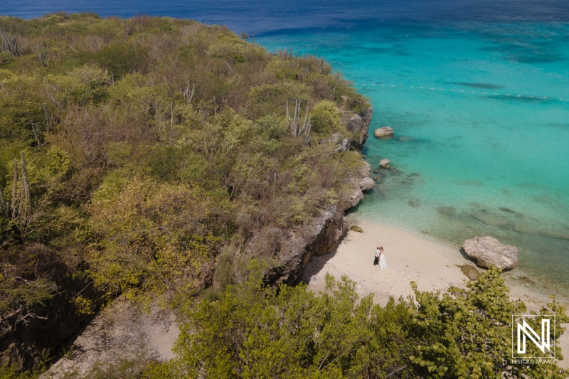 A couple stands on a secluded beach surrounded by lush greenery and clear turquoise waters under a bright sky on a sunny day