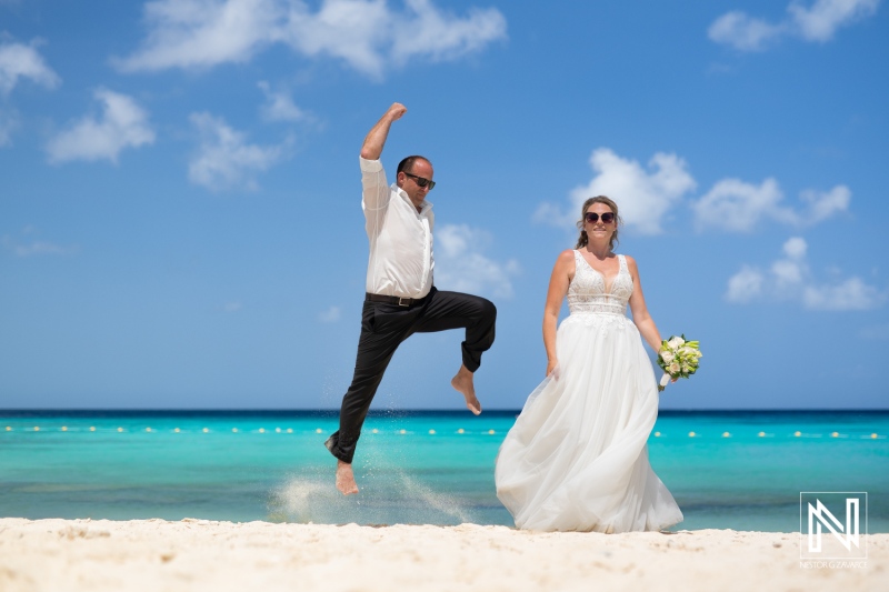 Bride in white dress and groom leaping on beach with clear turquoise water and blue sky, capturing joyful moment during wedding celebration in tropical paradise